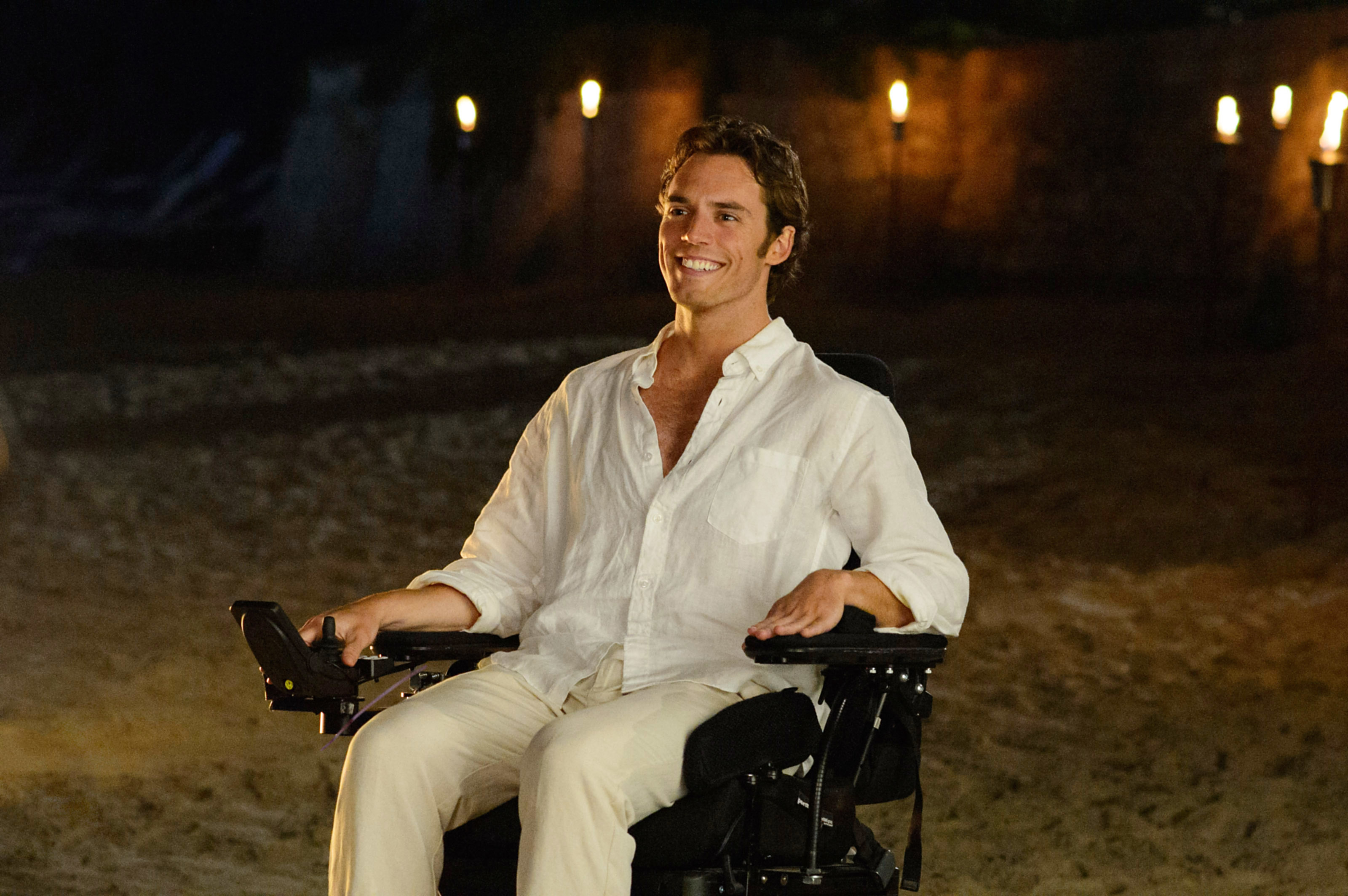 Sam Claflin in a white shirt and light pants, smiling, sits in a motorized wheelchair on a beach at night