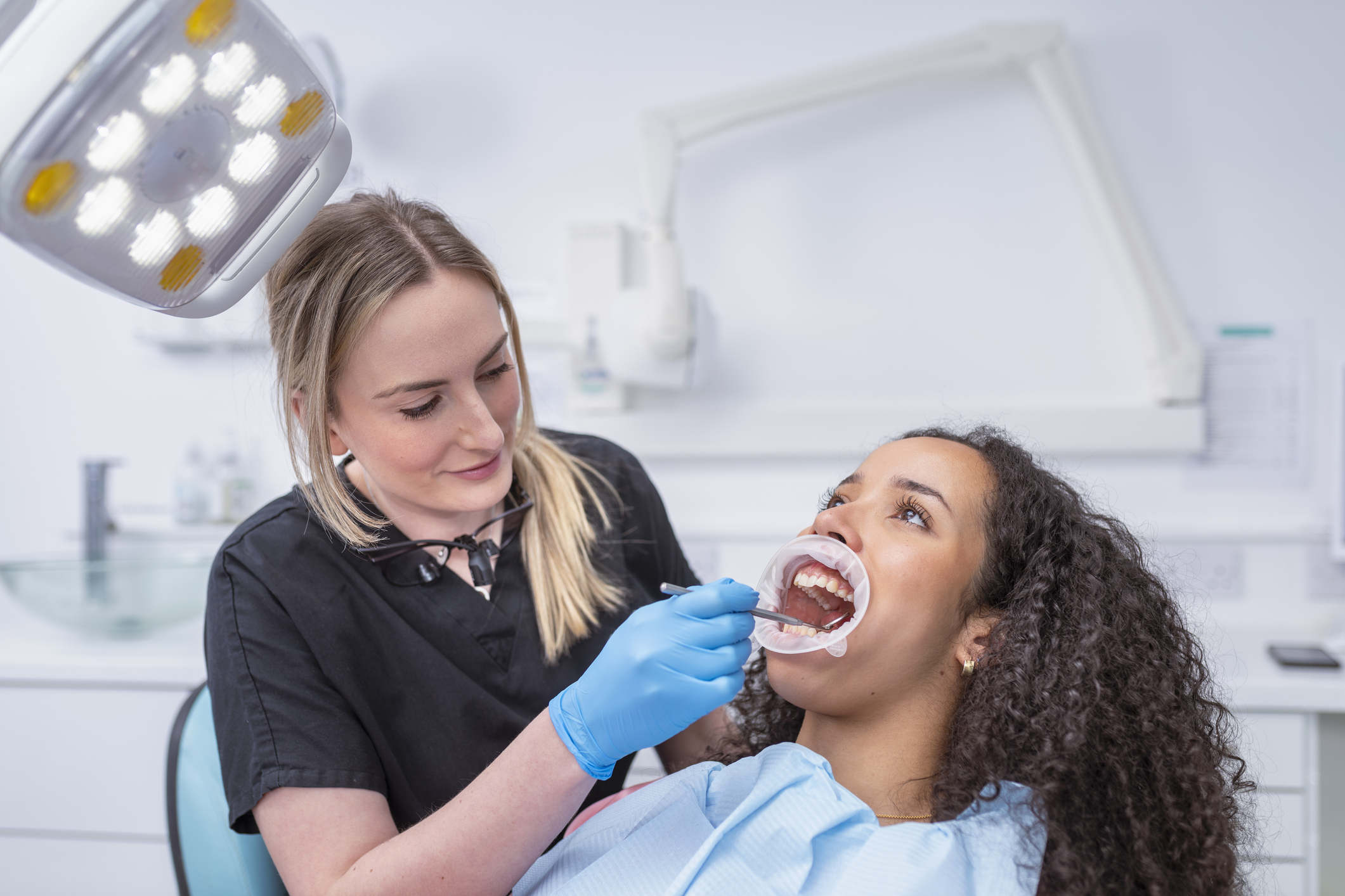 A female dentist in scrubs and gloves checks the teeth of a seated woman in a dental office