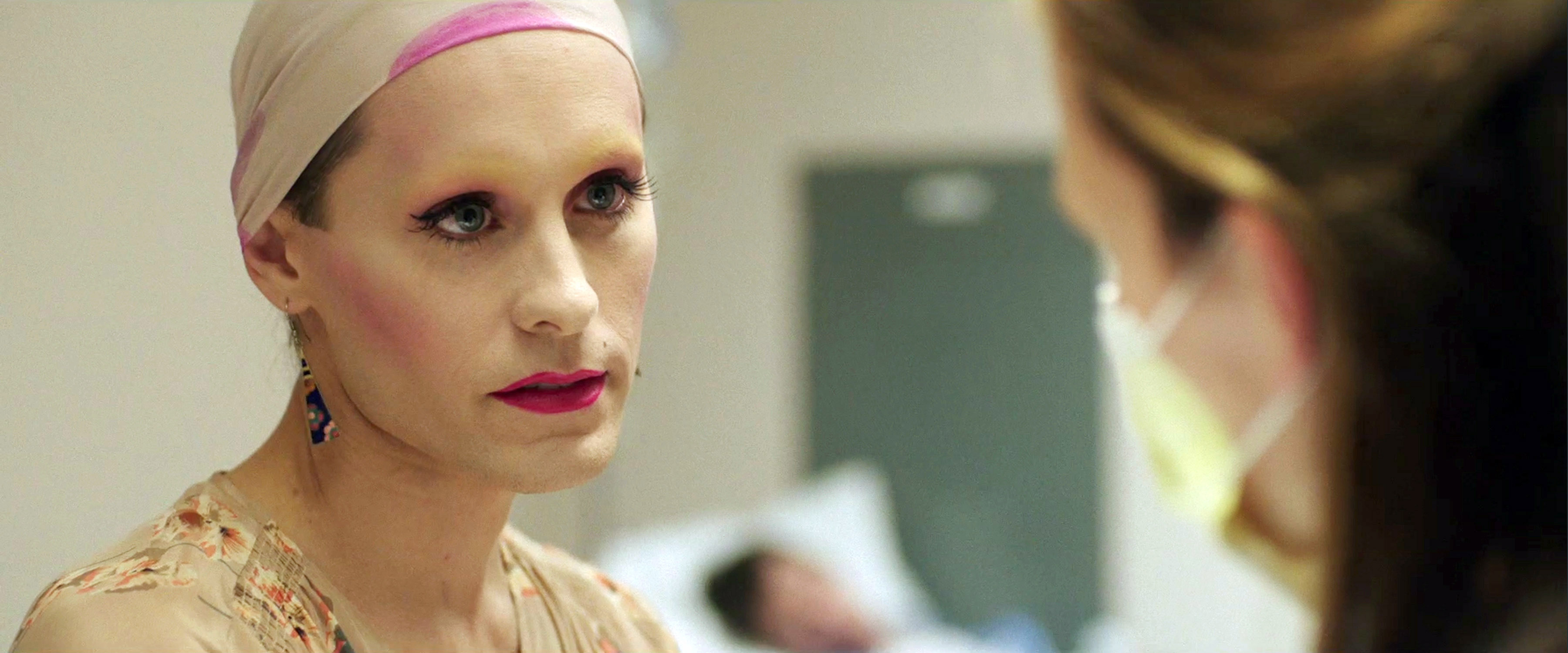 Jared Leto, portraying a character, wears a headscarf and makeup, speaking with a masked person in what appears to be a medical setting