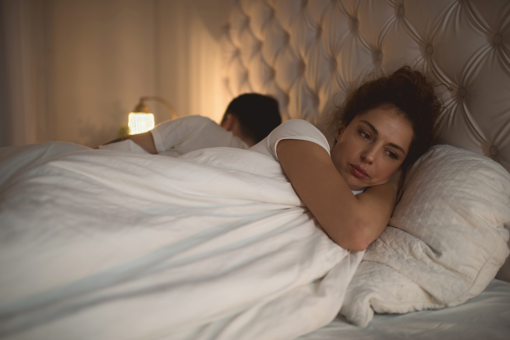 A woman, with a pensive expression, lies in bed next to a man who is turned away from her, suggesting a possible relationship issue