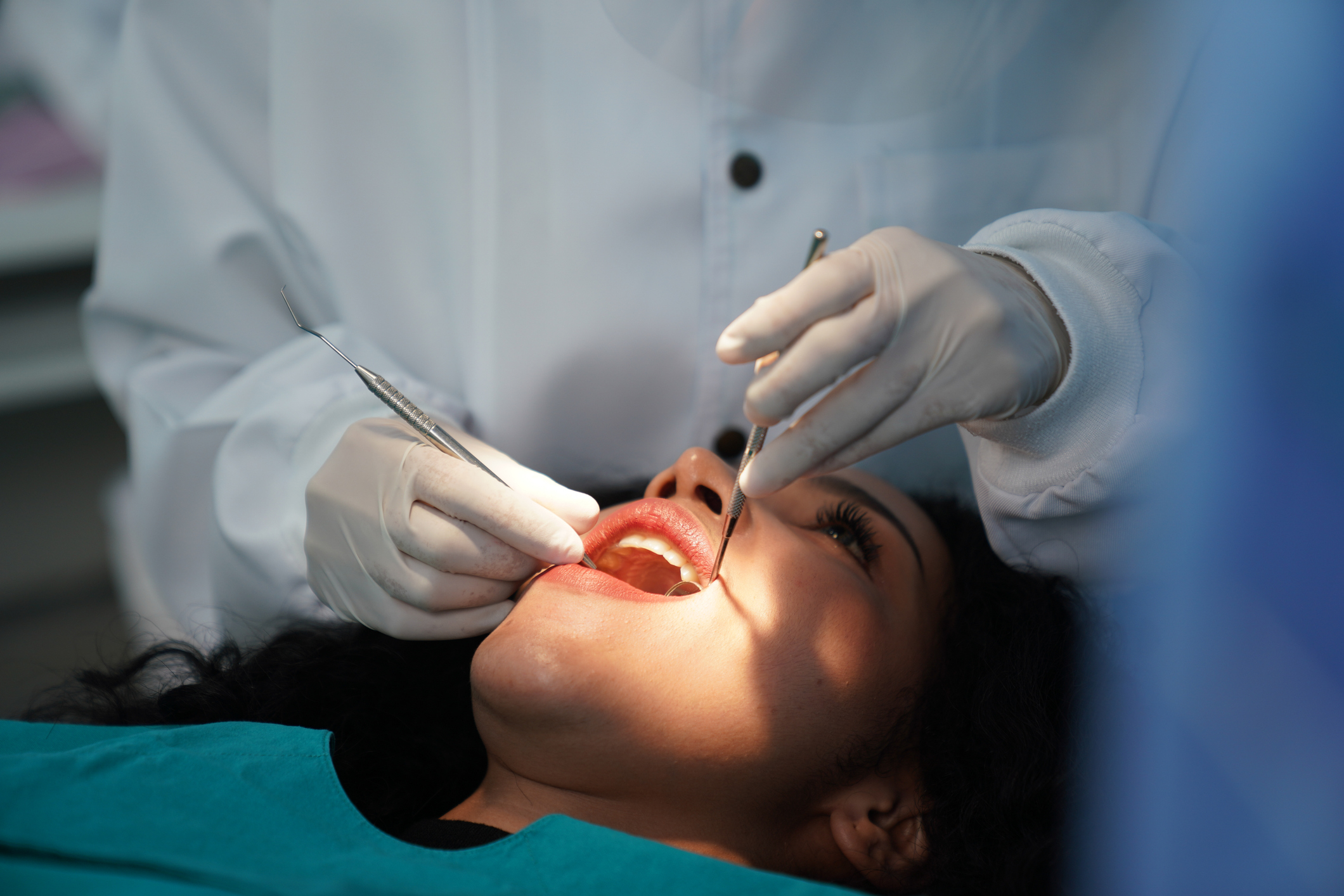 A patient gets a dental check-up from a dentist wearing white gloves and a lab coat, using dental tools while the patient lies back with their mouth open