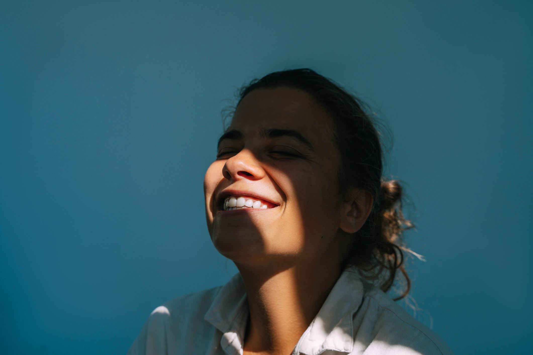 A woman with closed eyes smiles broadly, facing the sunlight. She is wearing a collared shirt