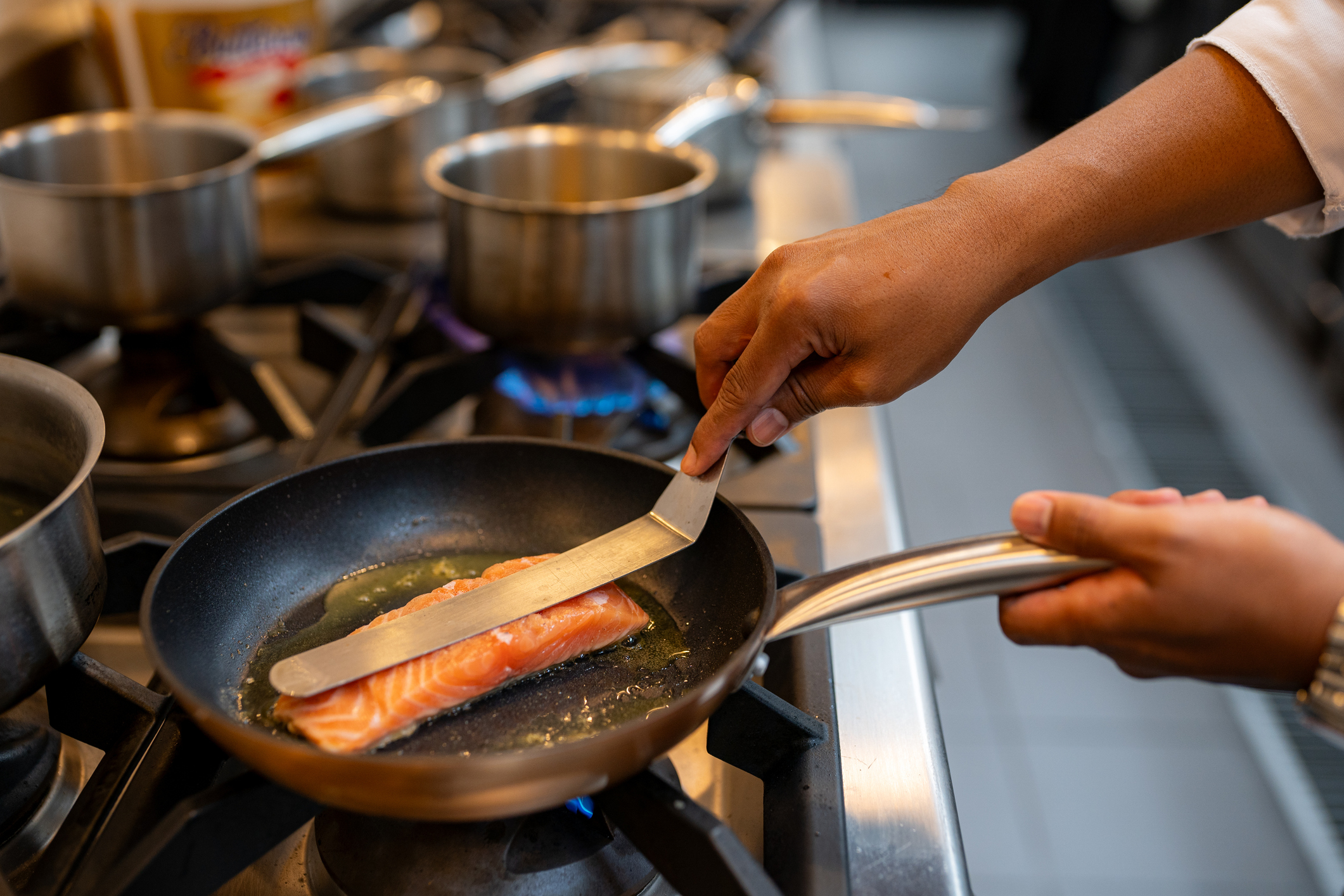 A person prepares a salmon fillet in a frying pan on a stove. Several pots are visible in the background