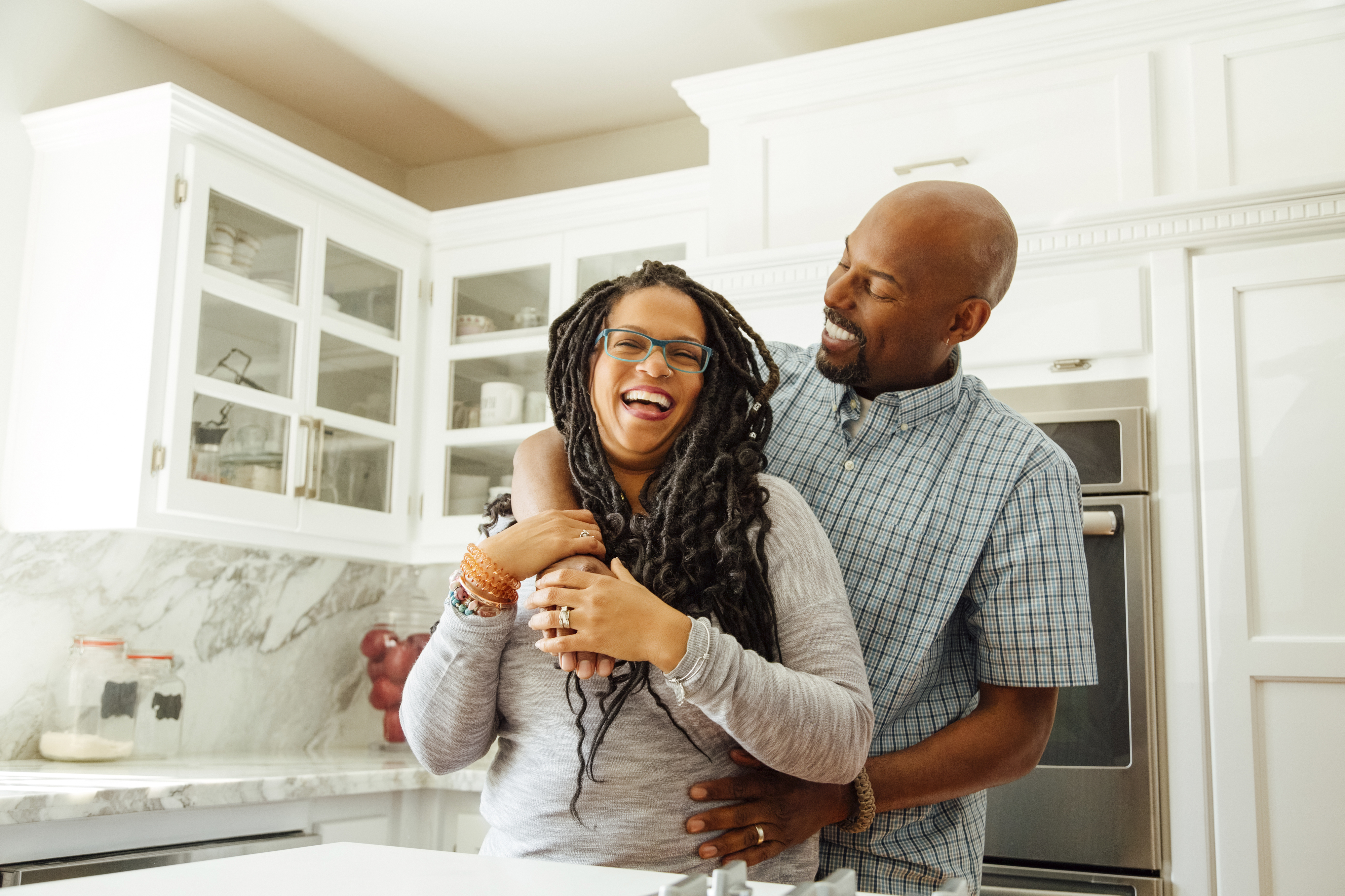 A woman and a man are standing in a kitchen. The man embraces the woman from behind, and they are both smiling and laughing
