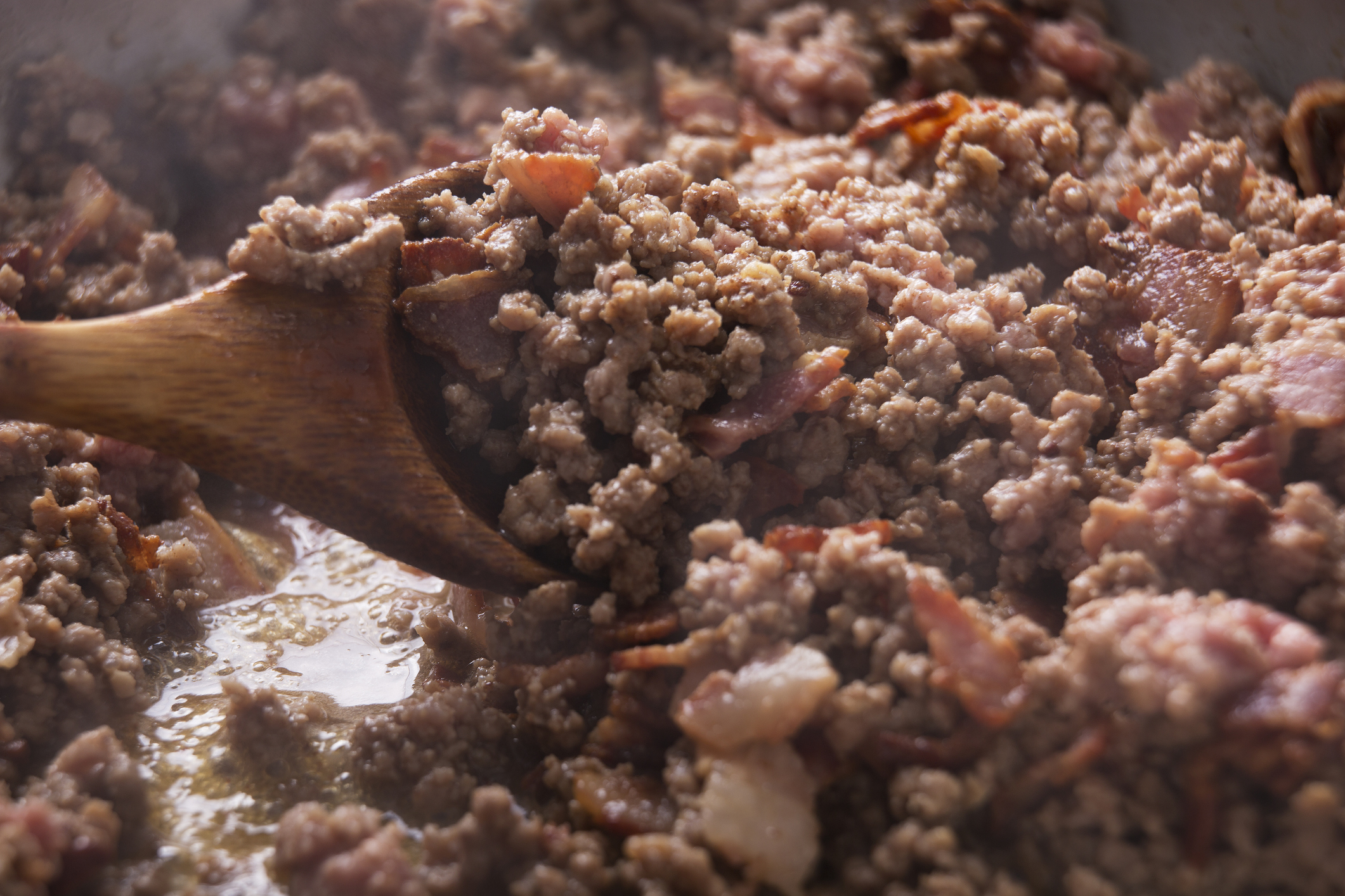 Ground meat and bacon are being stirred in a pan with a wooden spoon