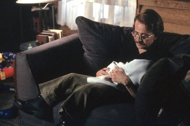 Man with glasses, in casual attire, sits on a sofa under a bright window, holding and gazing at a baby wrapped in a blanket