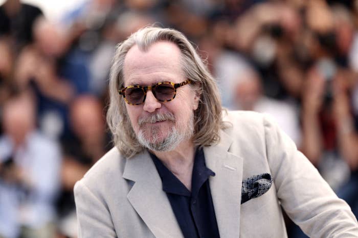 Gary Oldman wearing a beige jacket, navy shirt, and sunglasses, smiles at a public event, with photographers in the background
