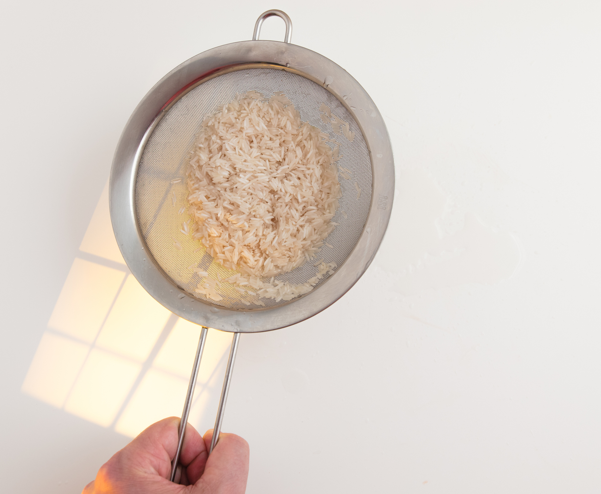 A person's hand holding a metal strainer filled with uncooked rice, draining in a kitchen setting