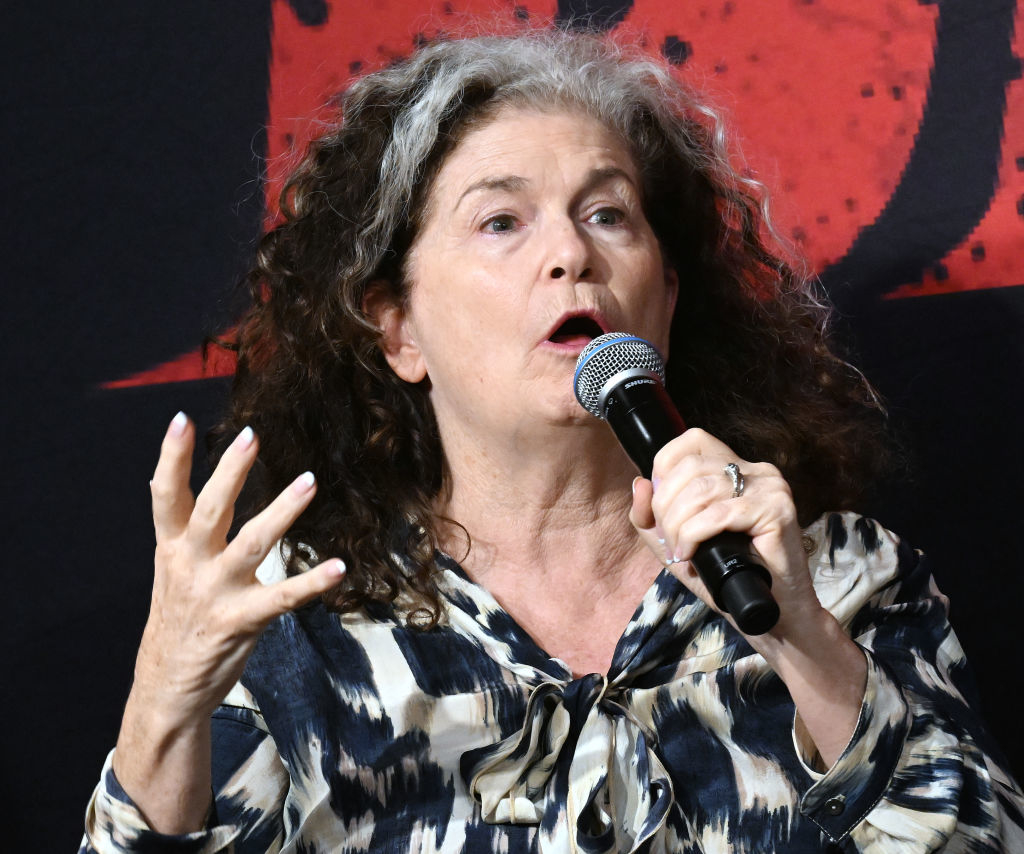 A woman with curly hair speaks passionately into a microphone while gesturing with her hand. She is wearing a patterned blouse