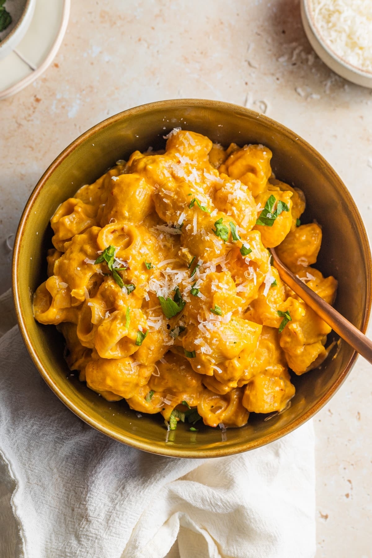 A bowl of creamy pasta topped with grated cheese and herbs, placed on a white cloth with a small bowl of grated cheese nearby