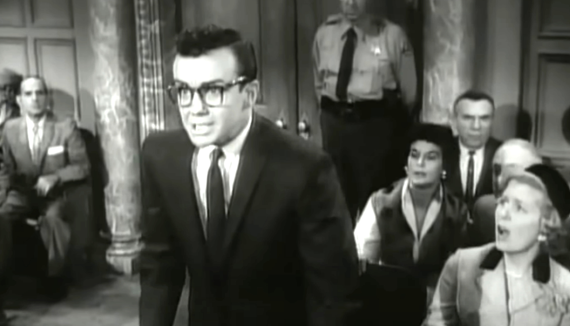 Courtroom scene with a man wearing glasses and a suit speaking passionately. Seated in the background are people including an officer and a woman in a hat