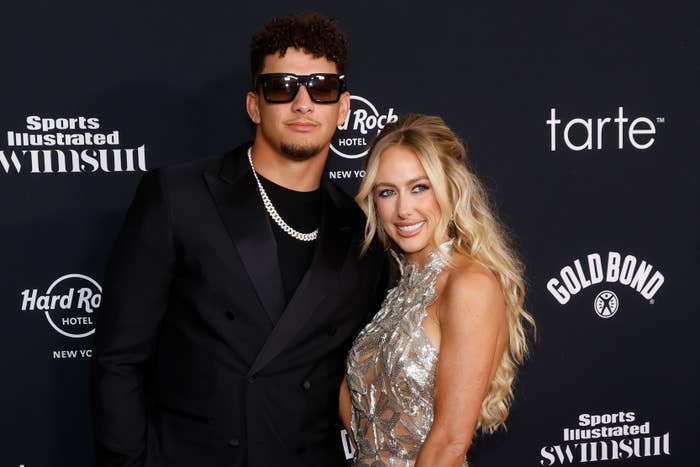 Patrick Mahomes in a dark suit with sunglasses and Brittany Mahomes in a sequined dress at a Sports Illustrated event