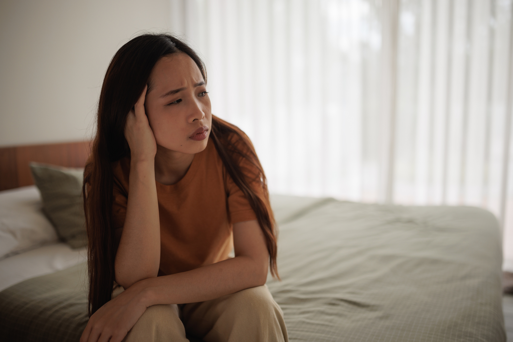 A woman sits on a bed with a concerned expression, resting her head on her hand