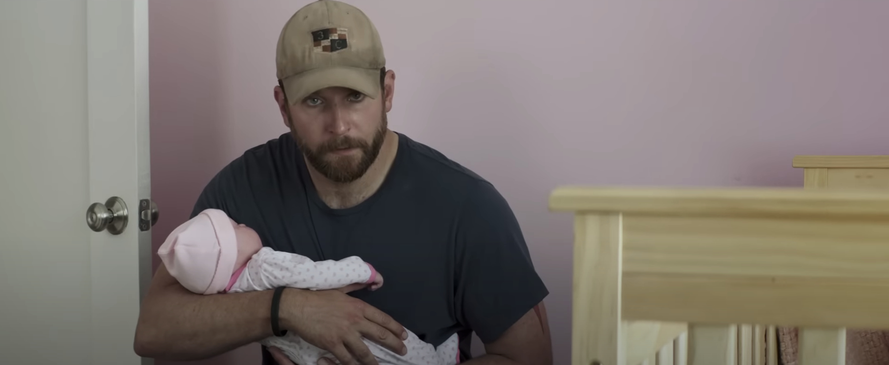 A man with a beard, wearing a cap, cradles a baby in his arms while sitting next to a crib in a baby&#x27;s room