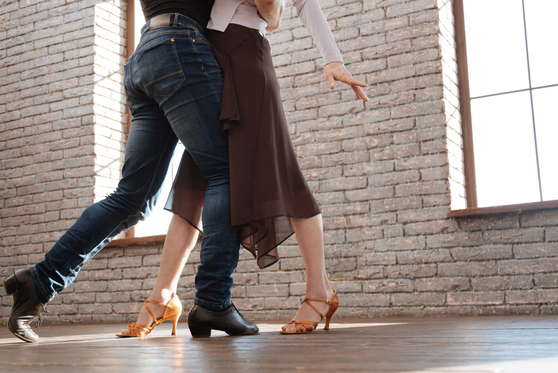 Two people dancing closely on a wooden floor in a spacious room with brick walls and large windows. Their faces are not visible