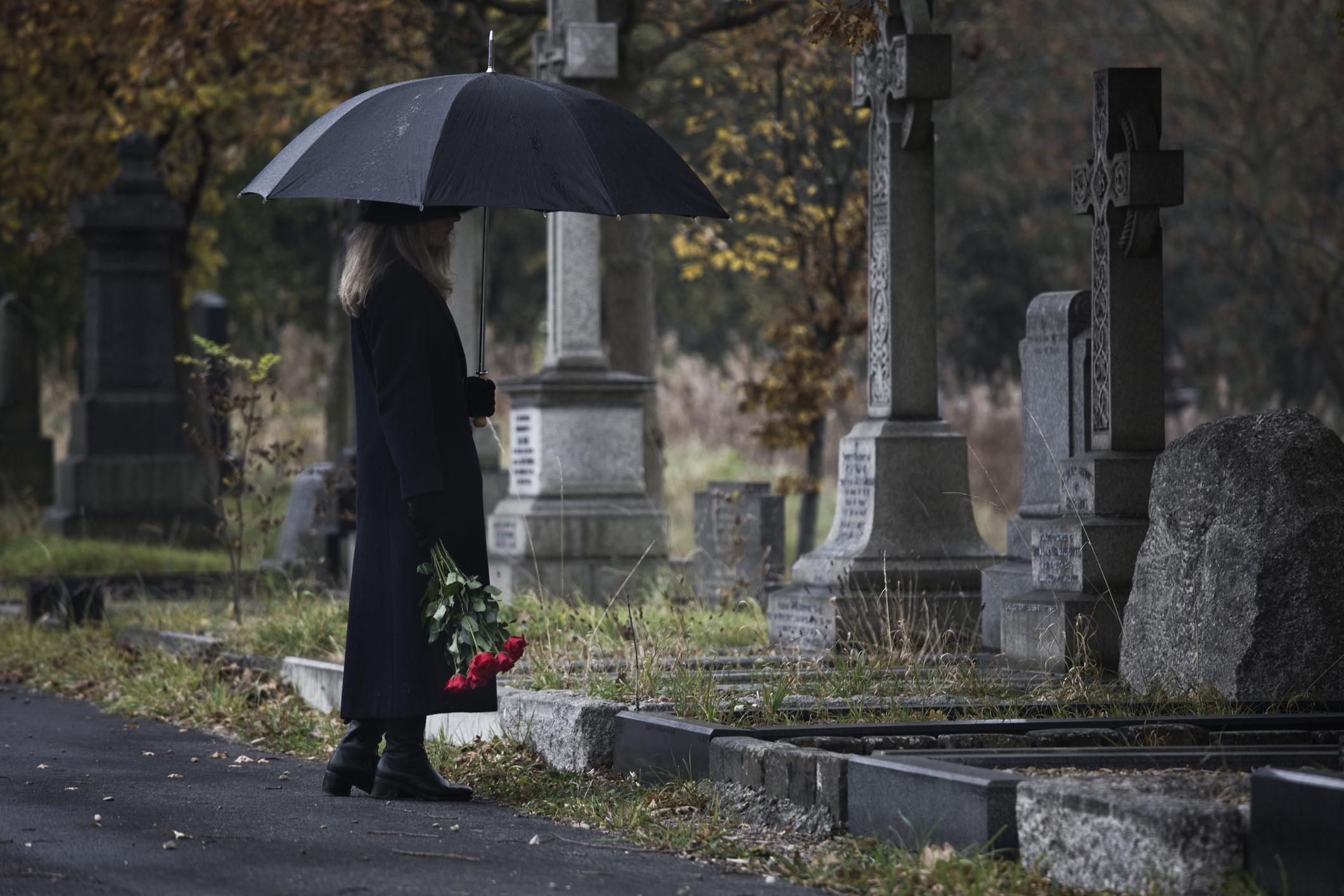 A person holding an umbrella and a bouquet of red flowers stands in front of a grave in a cemetery, surrounded by tombstones