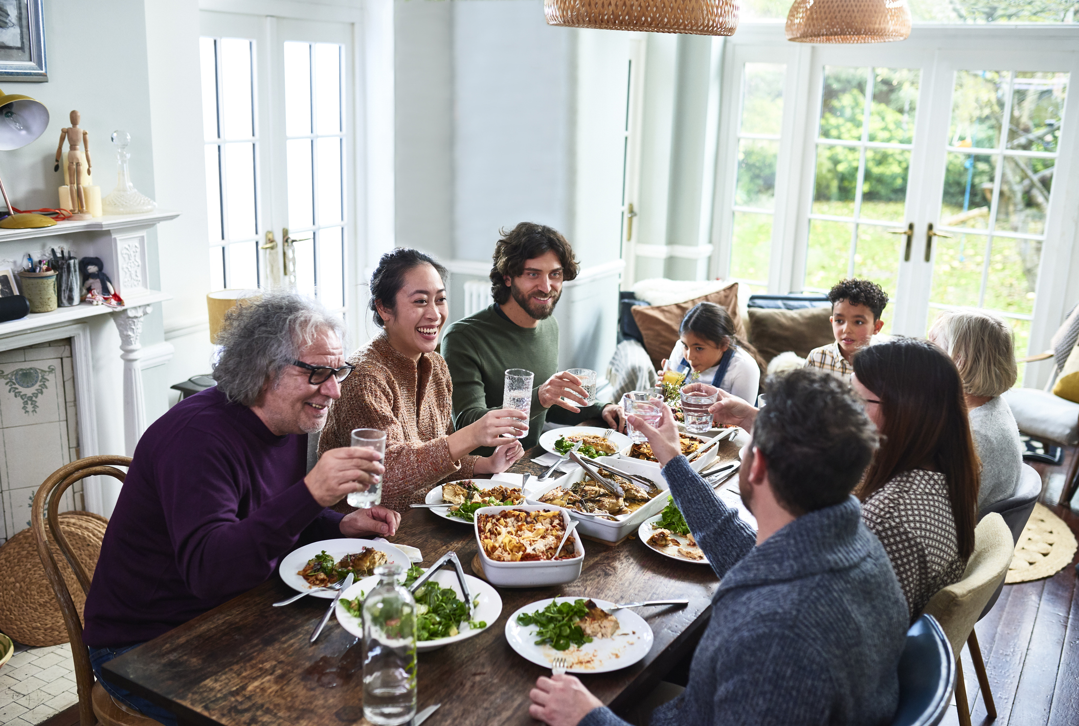 A family, including three adults and three children, enjoys a meal together at a dining table at home, smiling and raising glasses in a toast