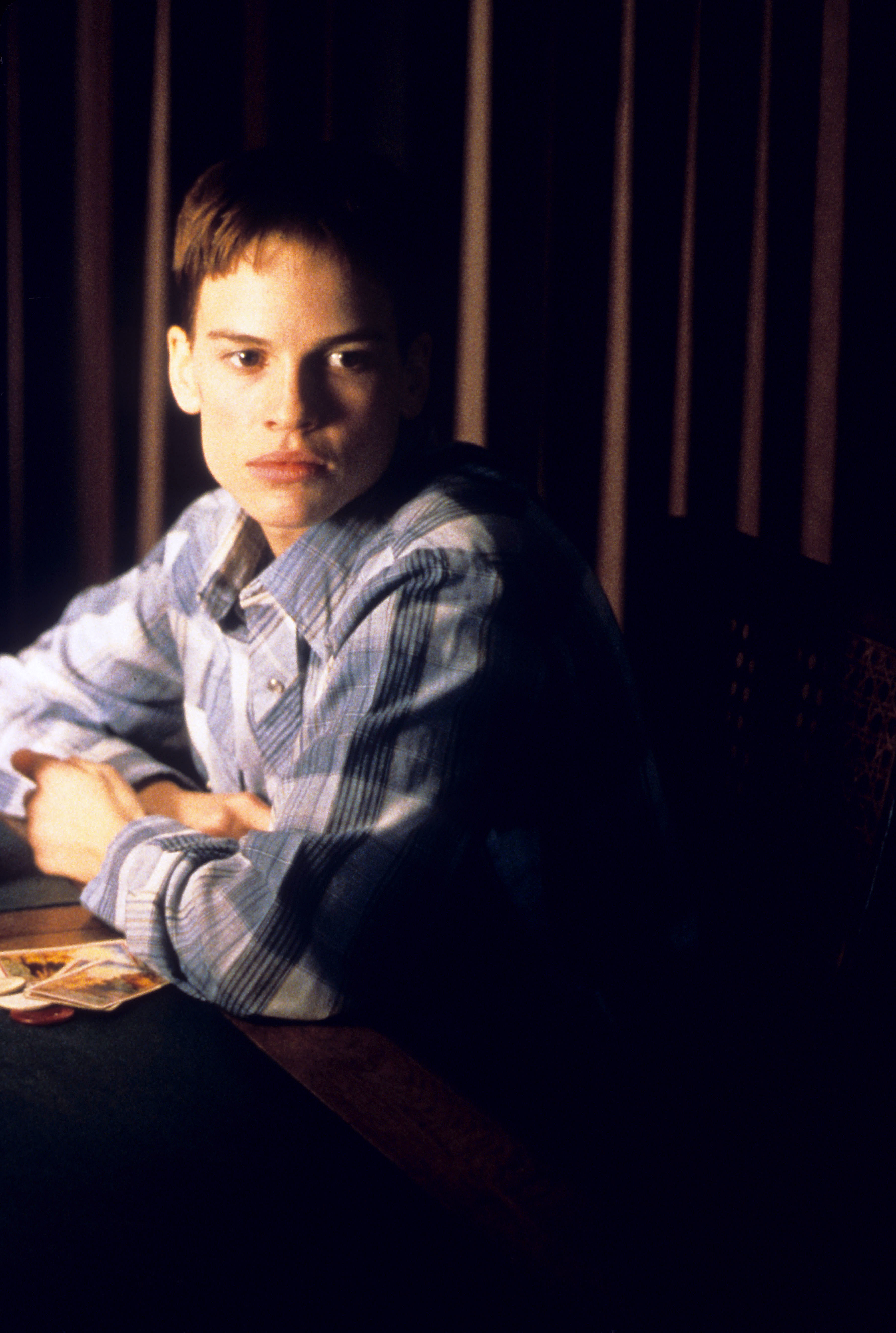 Hilary Swank sits at a table with playing cards in a casual plaid shirt, looking off to the side