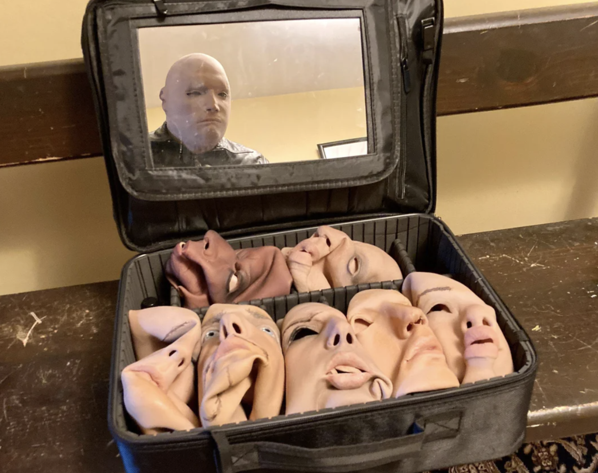 A person with a shaven head is reflected in a mirror on an open case filled with various realistic face masks