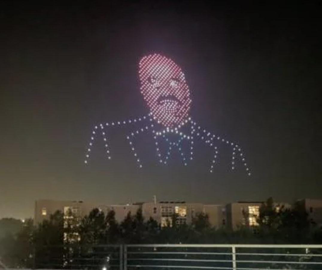 A large formation of drones in the night sky creates a light display of a man's face wearing a suit and bow tie. Trees and buildings are visible below