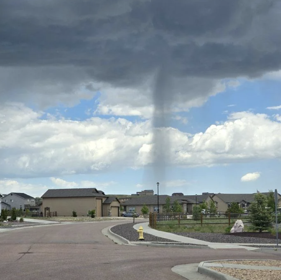 A funnel cloud forming over a suburban residential area, with houses and a fire hydrant visible in the foreground