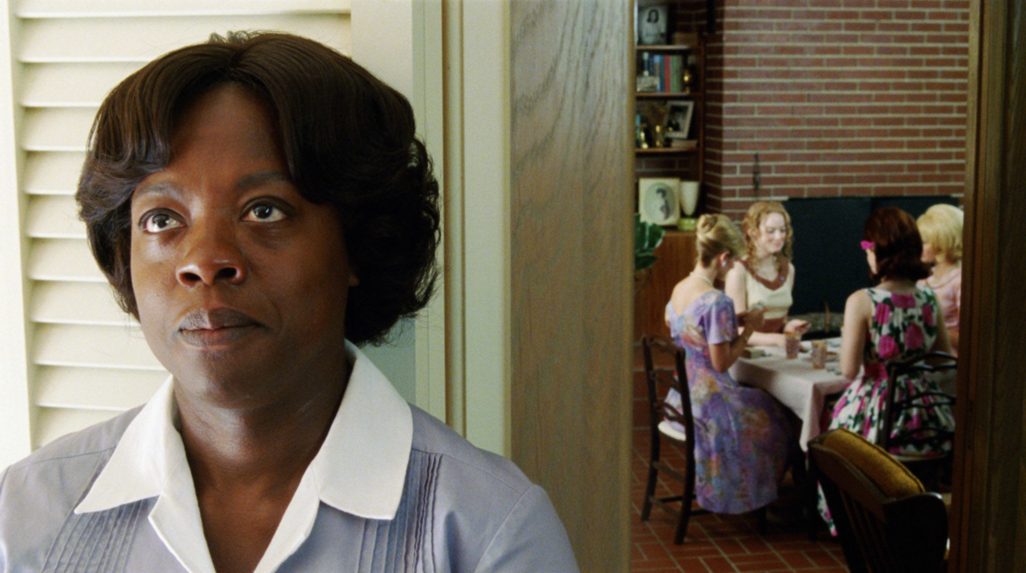 Viola Davis stands in the foreground looking serious; in the background, four women sit at a table in discussion