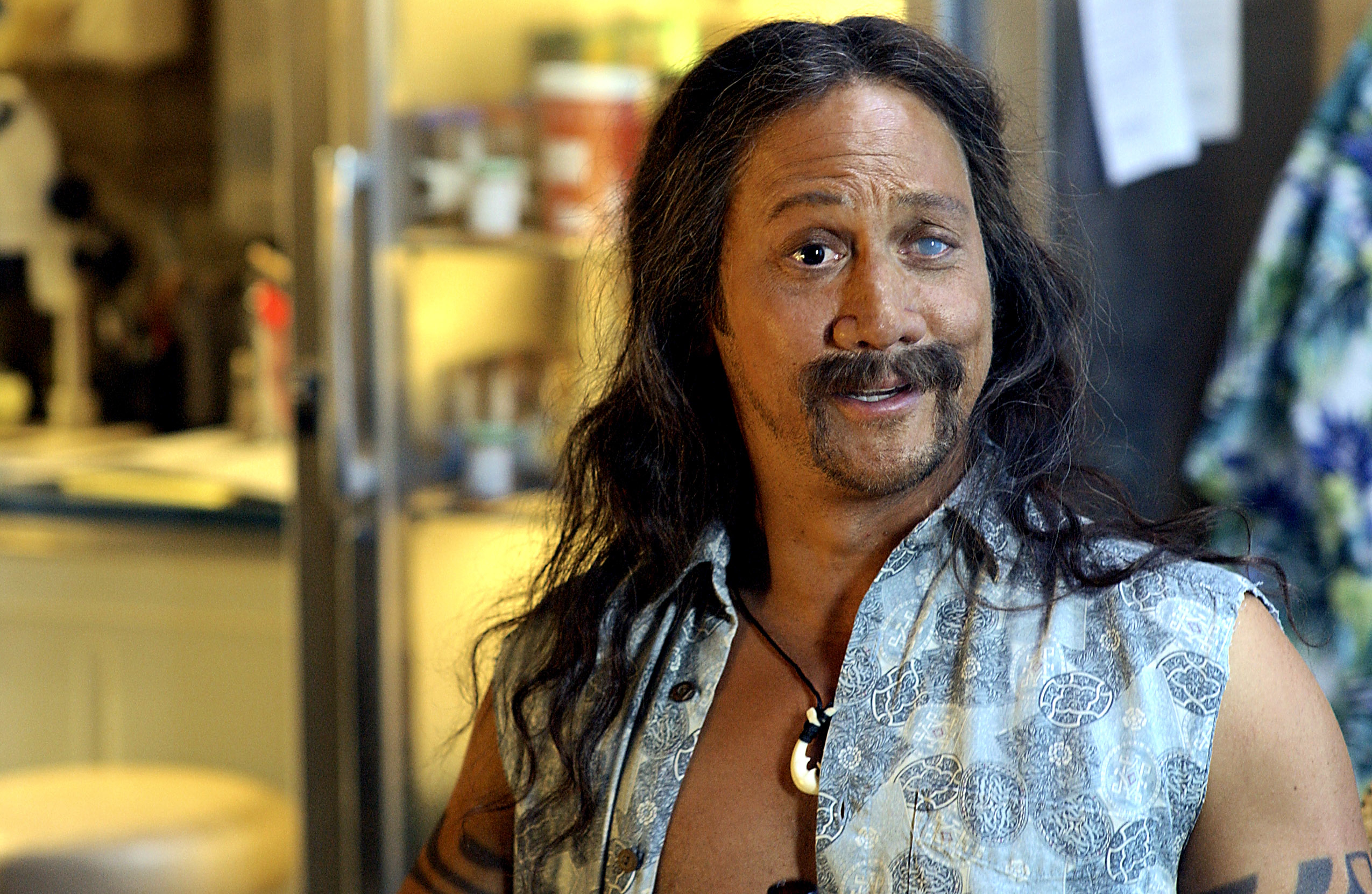 Man with long hair, mustache, and goatee, wearing an open sleeveless shirt, smiles in a kitchen setting