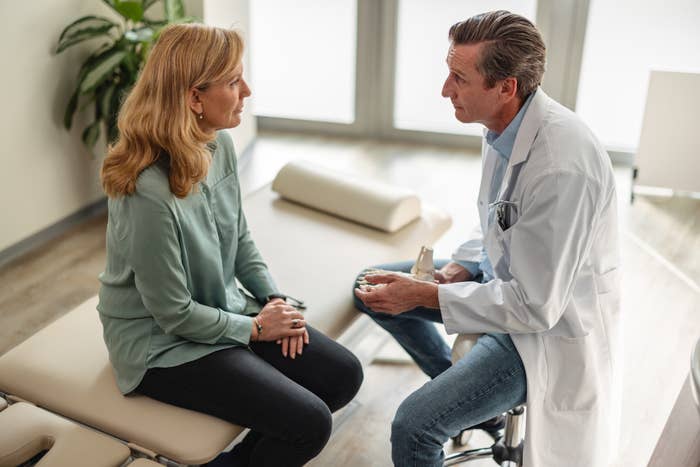 A doctor, wearing a white lab coat, speaks with a woman in casual attire during a consultation in a medical office. Both are seated and engaged in conversation
