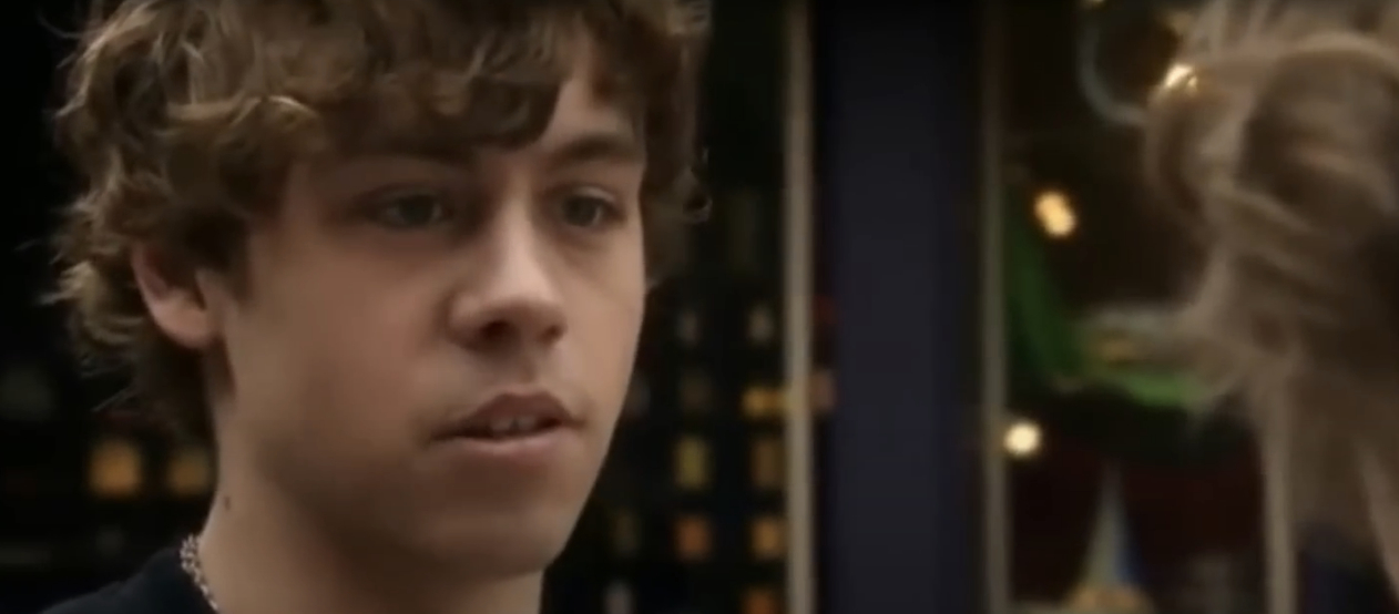 A young man with curly hair looks concerned while talking to another person in a close-up scene from a movie or TV show. Names of persons unknown