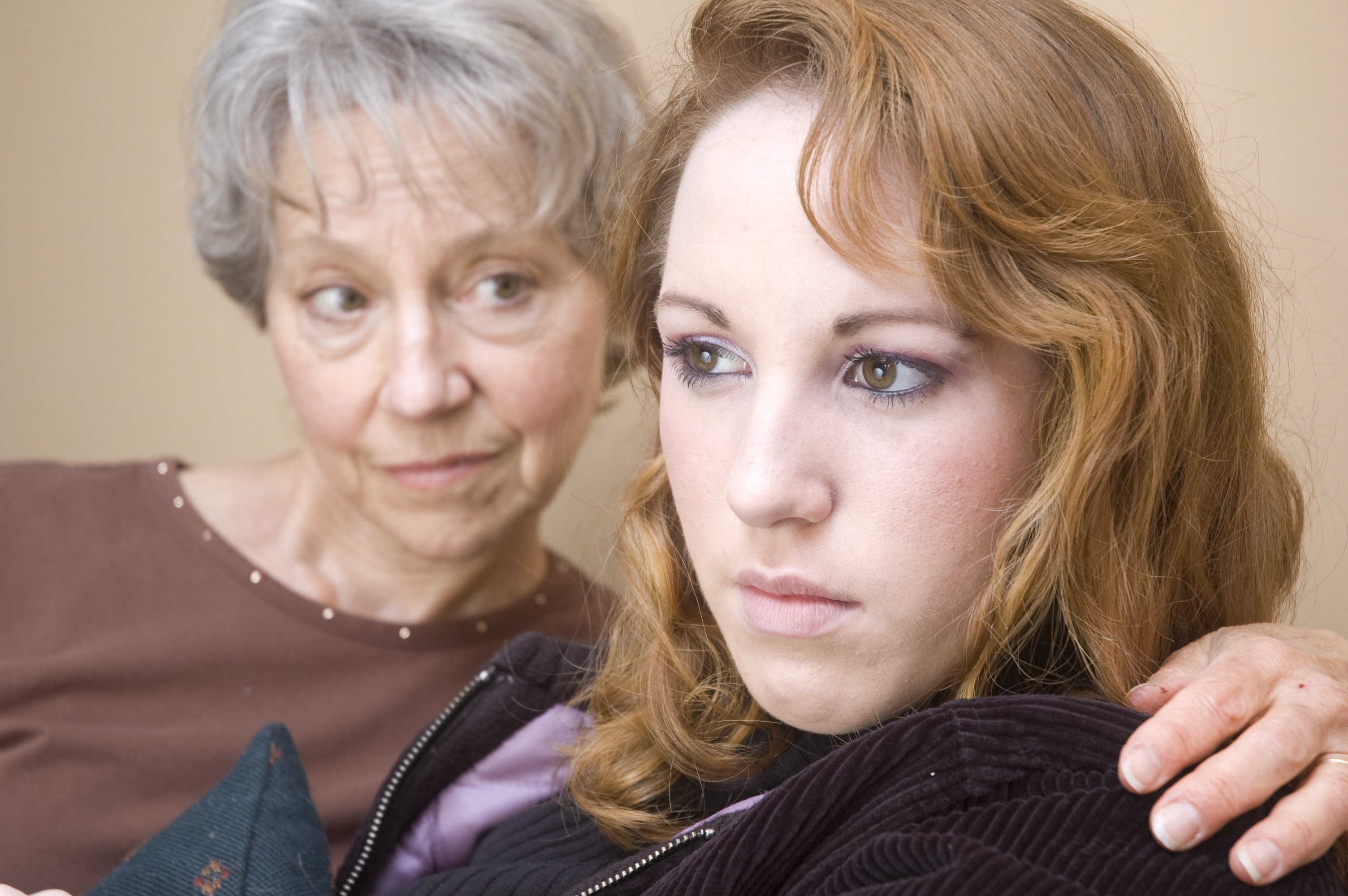 An older woman with short, gray hair looks at a younger woman with long, light hair who appears upset. The older woman has her hand on the younger woman's shoulder