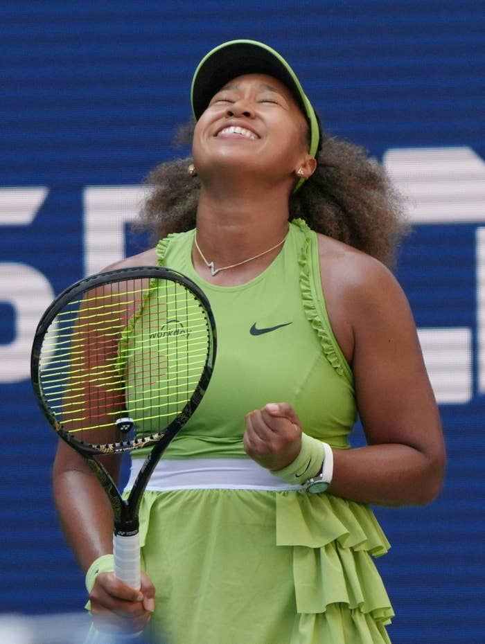 Naomi Osaka, wearing athletic tennis attire, clenches her fist and smiles towards the sky on a tennis court