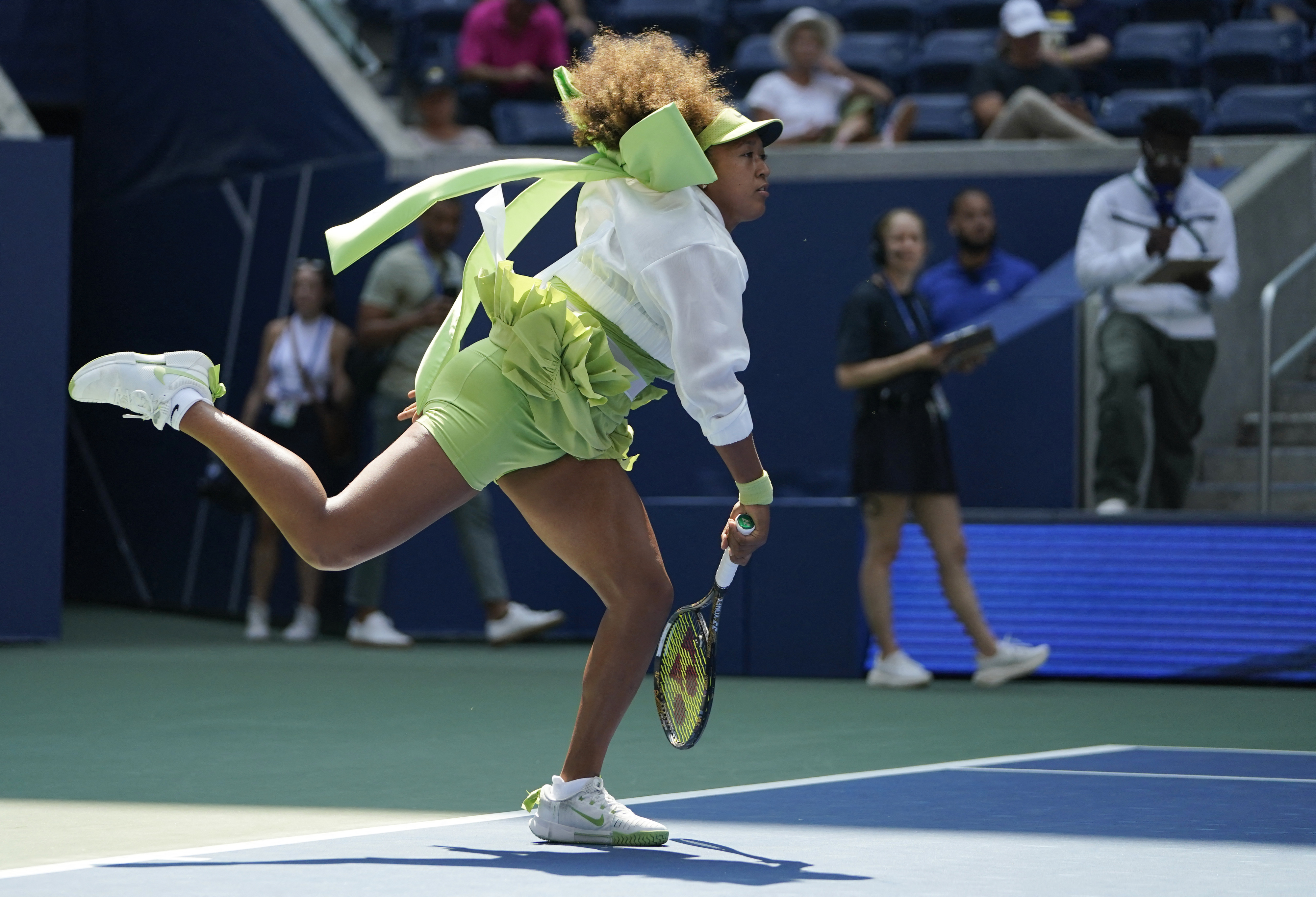 Naomi Osaka in mid-action during a tennis match, wearing a sporty outfit with a large bow on her back. She is focused and poised, ready to hit the ball