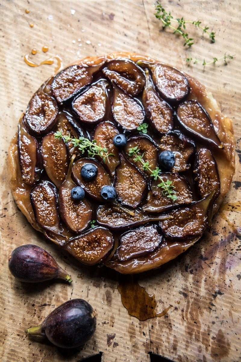 A rustic fig tart decorated with fresh figs, blueberries, and thyme, on a textured surface with additional figs nearby