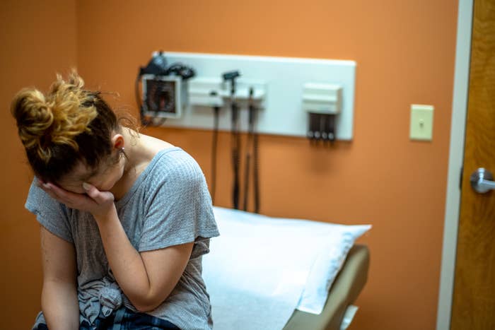 A woman sits on an exam table in a doctor's office, covering her face with one hand, appearing distressed. Medical equipment is visible on the wall behind her