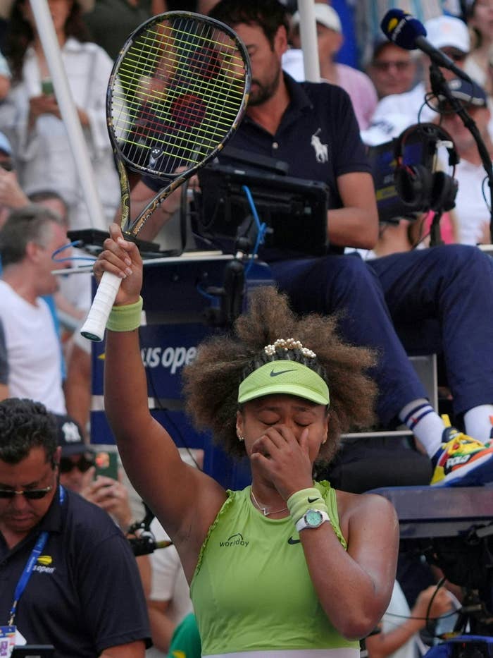 Naomi Osaka on a tennis court, holding a racket up and covering her mouth with her other hand, showing an emotional reaction