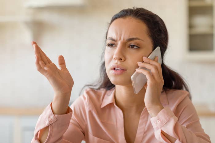 A woman with long dark hair, wearing a buttoned shirt, is talking on her phone with a frustrated expression, gesturing with her other hand