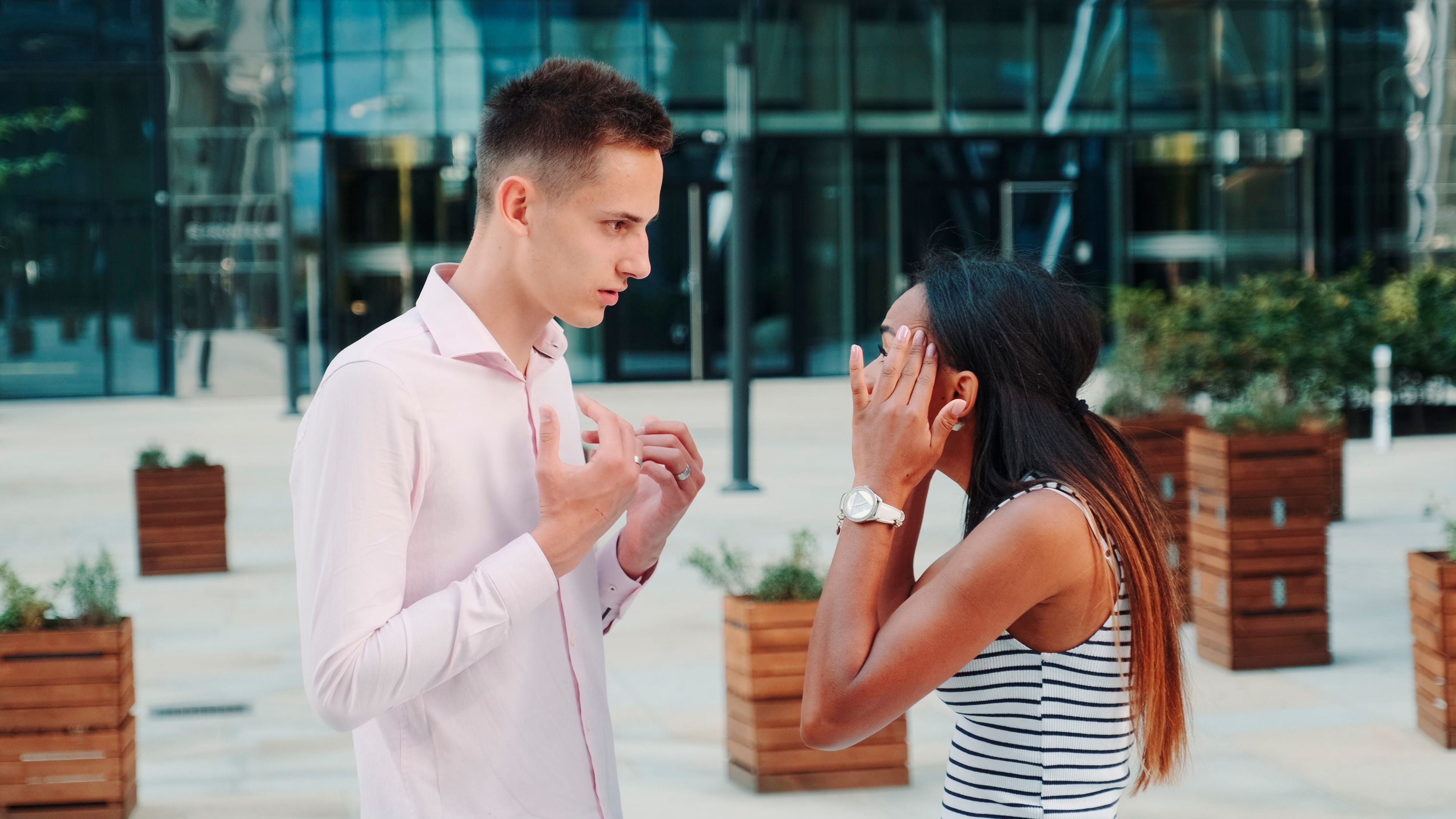A man and woman are having an intense conversation outside a modern building. The man looks upset, while the woman has her hands on her head, looking stressed