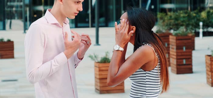 A man and woman are having an intense conversation outside a modern building. The man looks upset, while the woman has her hands on her head, looking stressed