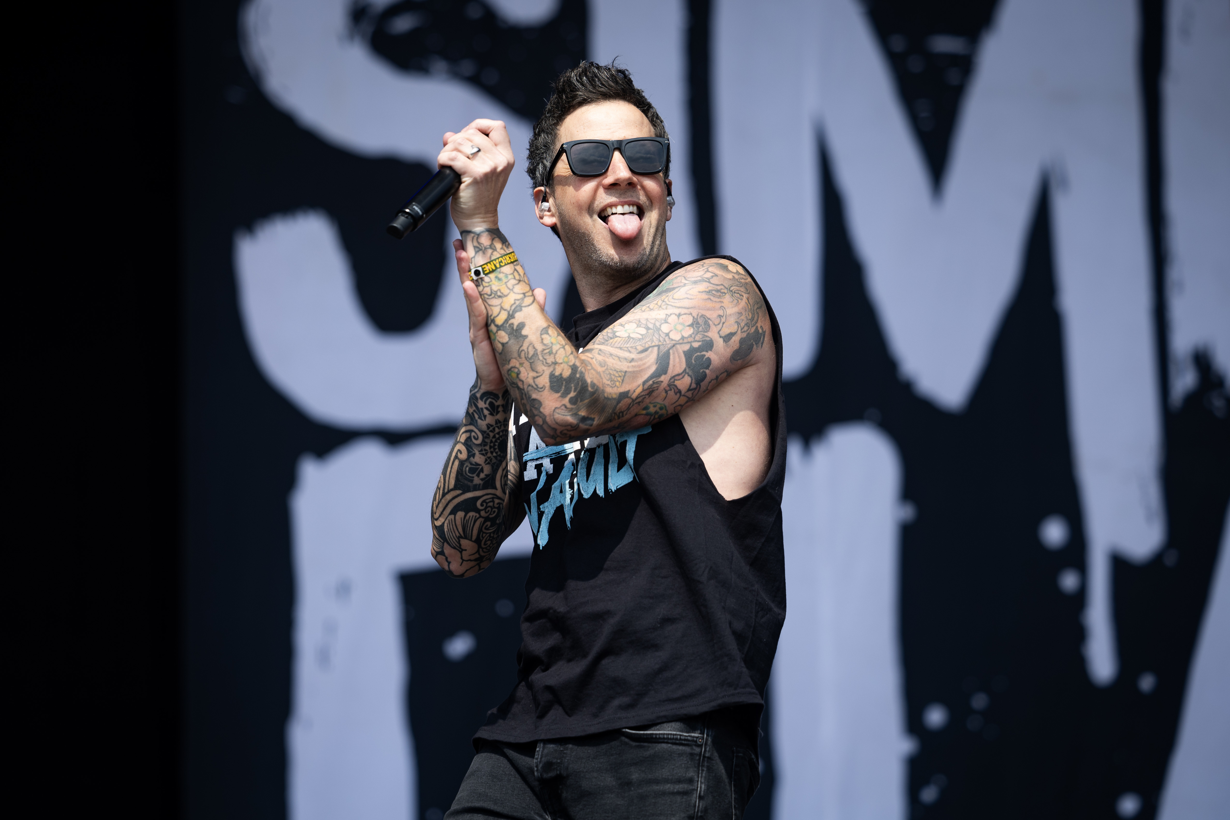 Pierre Bouvier of Simple Plan, wearing sunglasses, a sleeveless shirt, and showcasing tattoos, performs energetically on stage, microphone in hand