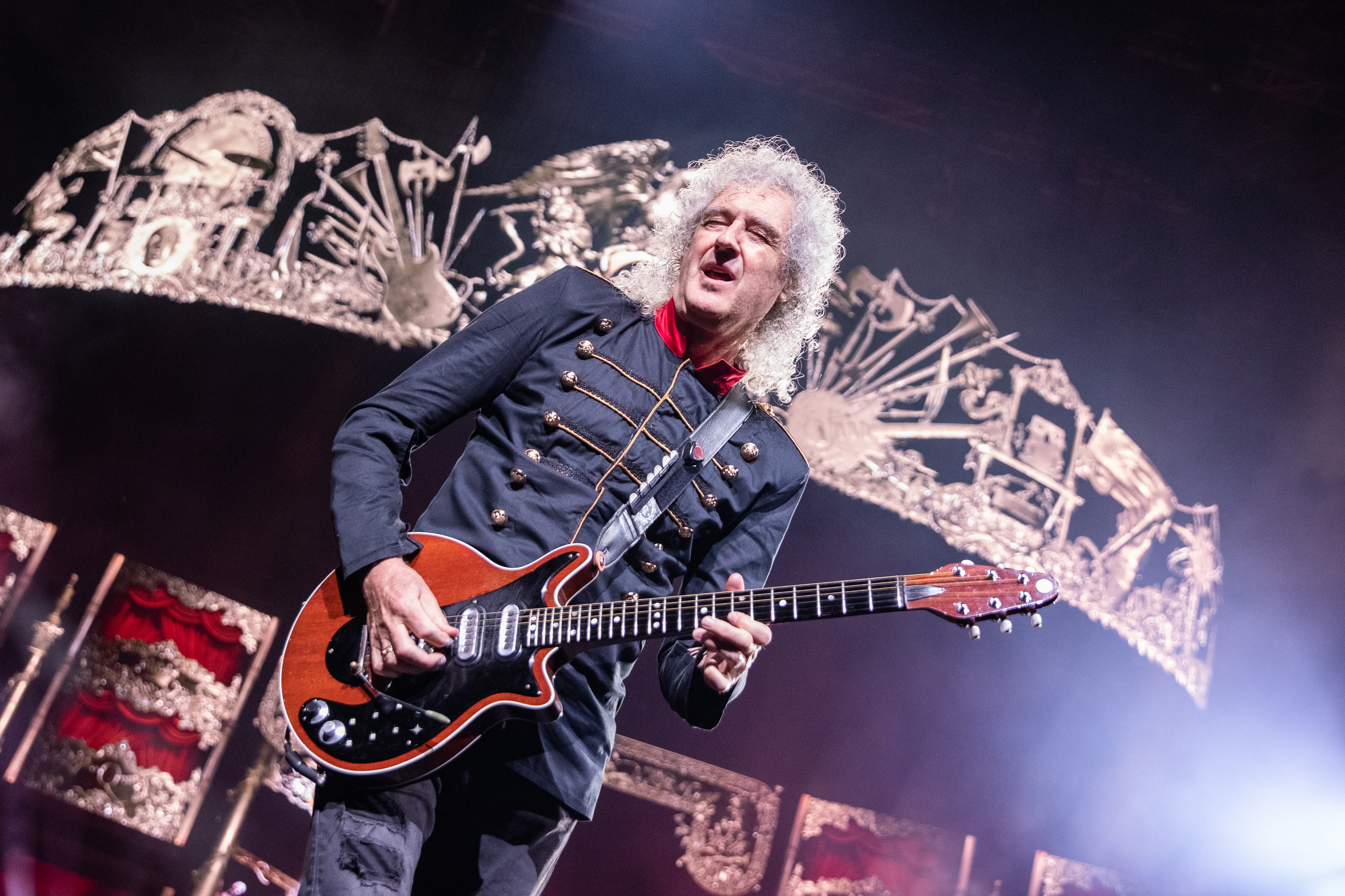 Brian May performs on stage, playing guitar, dressed in a military-style jacket, with intricate stage decorations in the background