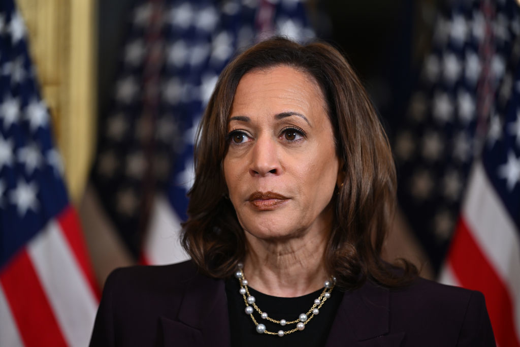 Kamala Harris is seen wearing a dark suit jacket and a pearl necklace, standing in front of American flags