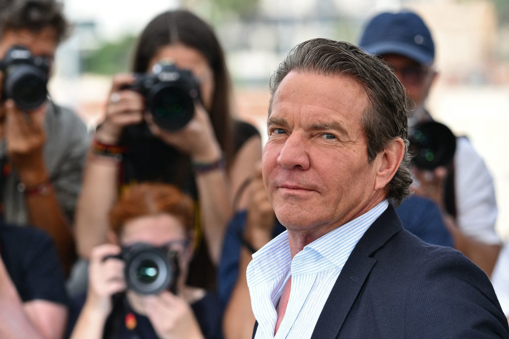 Dennis Quaid is standing confidently in front of photographers, wearing a dark suit jacket and a light shirt, sporting a slight smile
