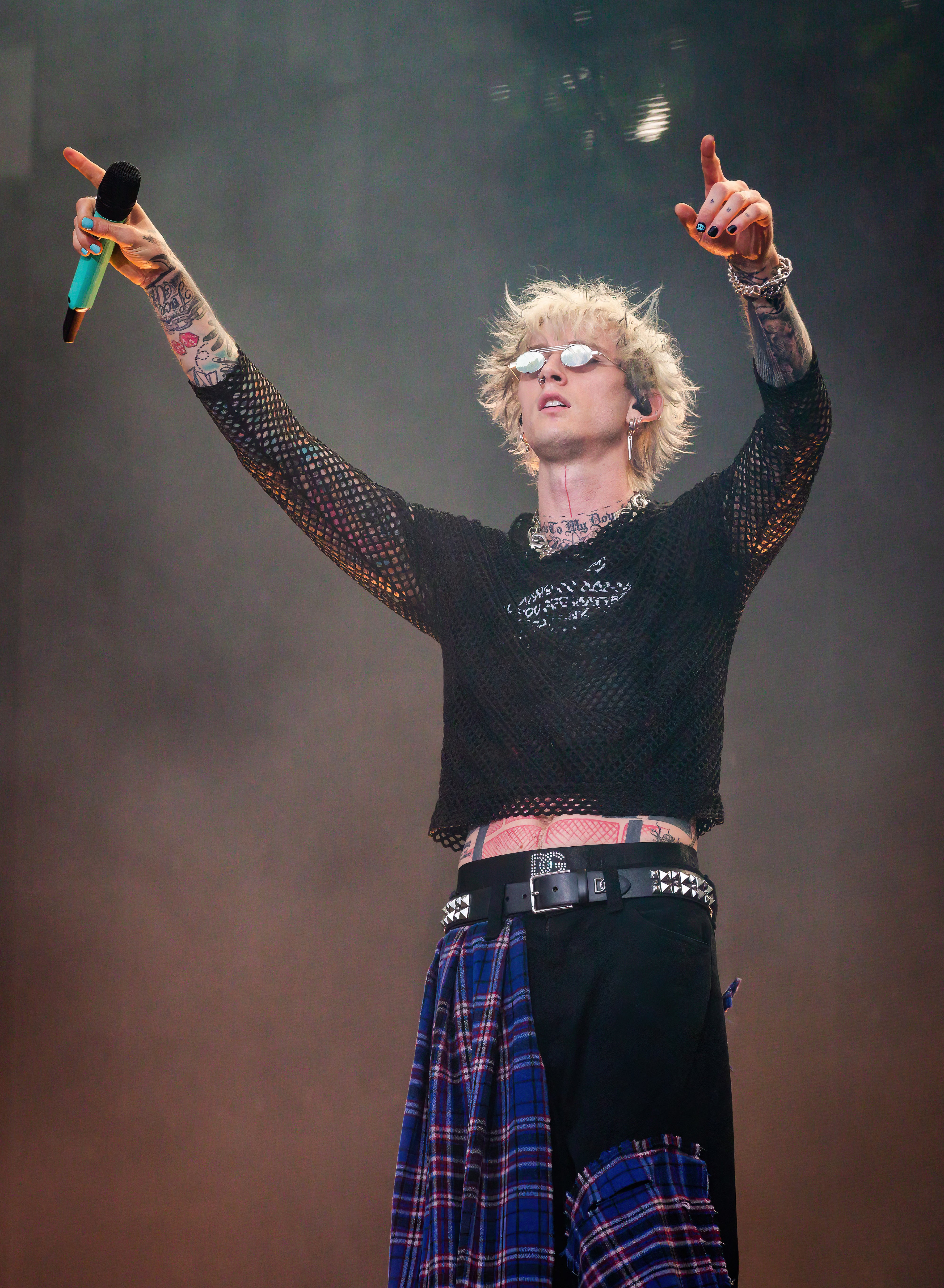 Machine Gun Kelly performs on stage, wearing a mesh shirt with a plaid flannel tied around his waist, holding a microphone and gesturing to the crowd