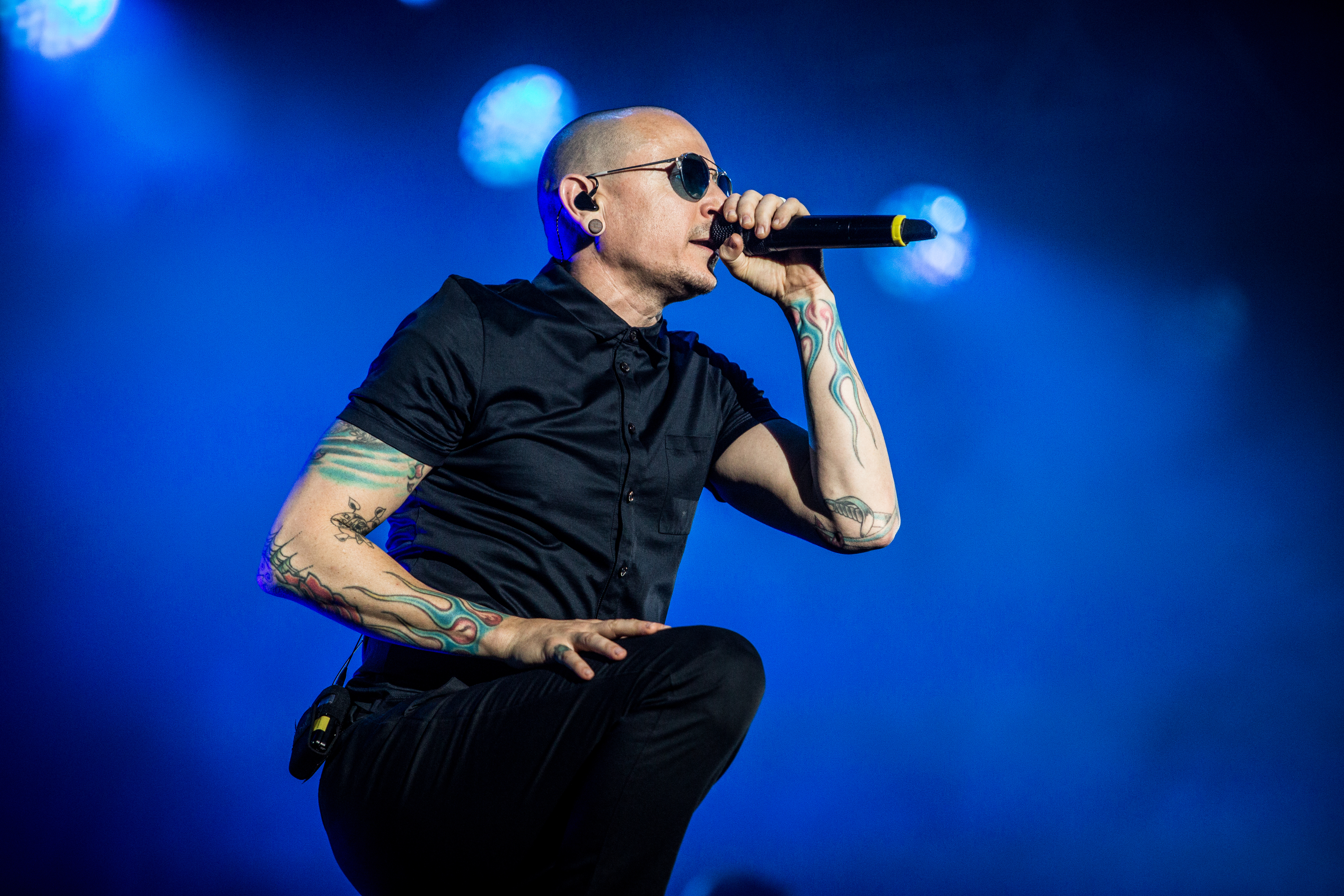 Chester Bennington is performing on stage, wearing sunglasses and an all-black outfit with colorful tattoos visible on his arms, in front of a blue background