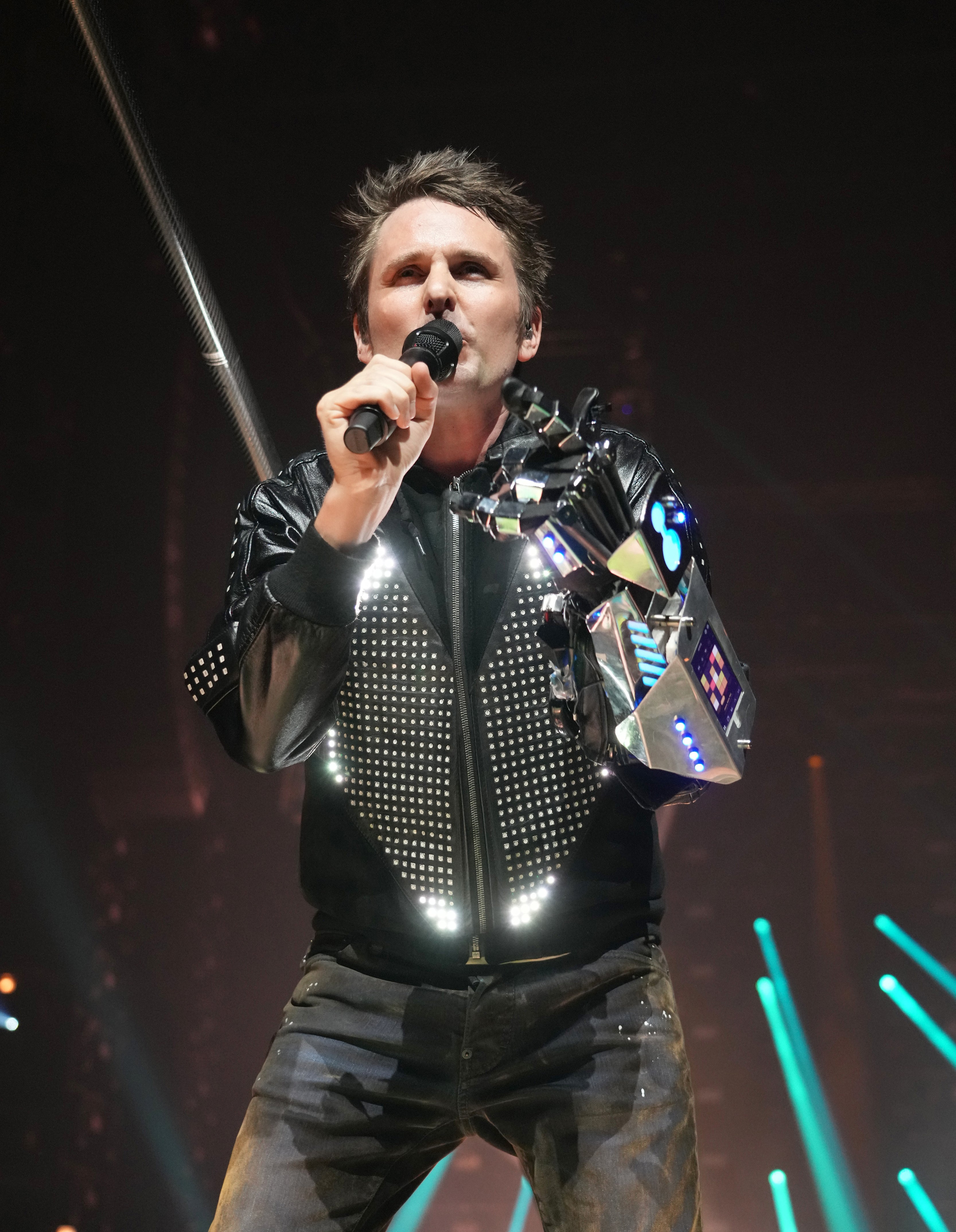 Matthew Bellamy, performing on stage, wearing a studded jacket and a futuristic, metallic arm accessory, holding a microphone