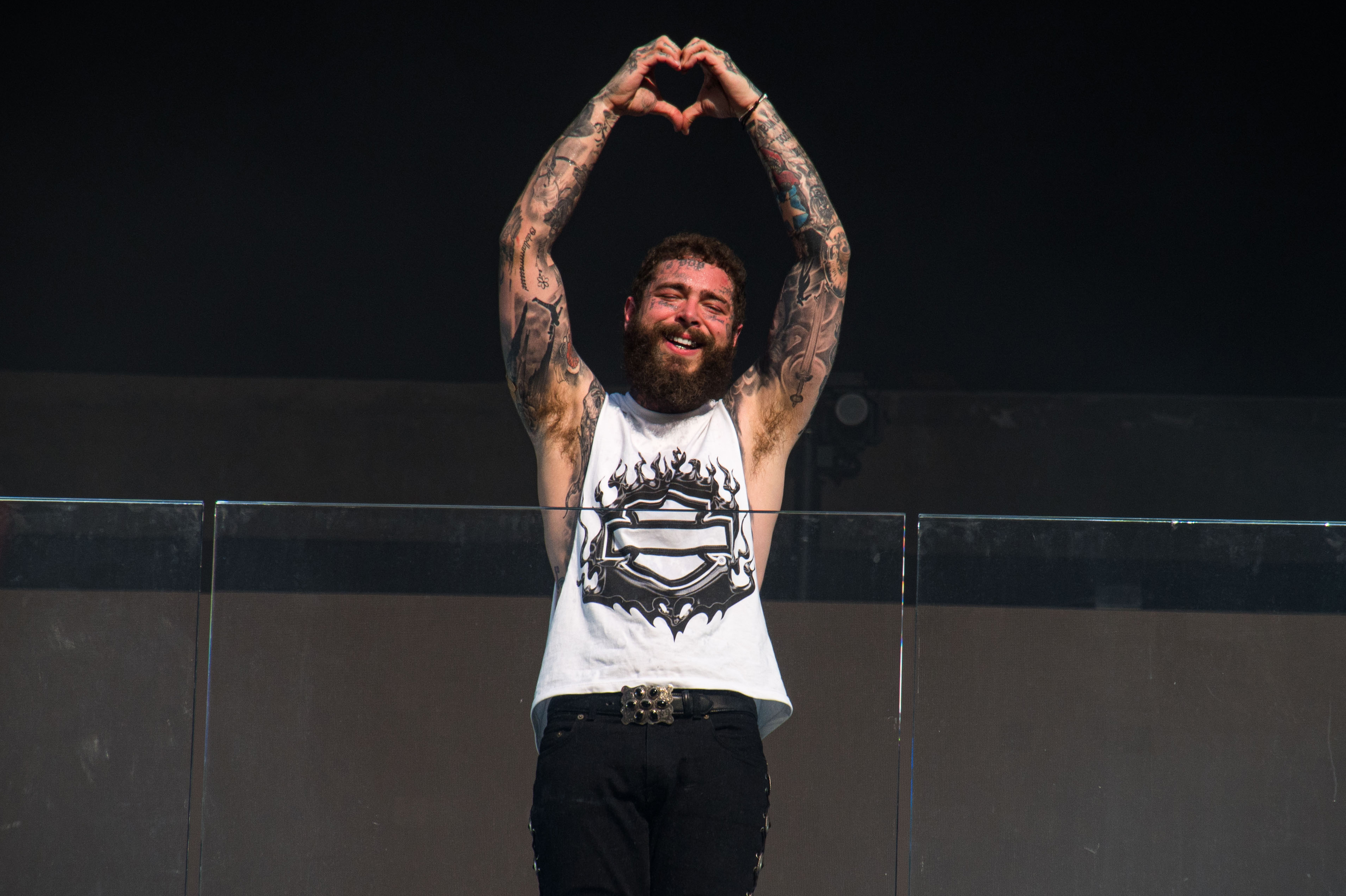 Post Malone wearing a sleeveless shirt with a graphic design, making a heart shape with his hands above his head on stage