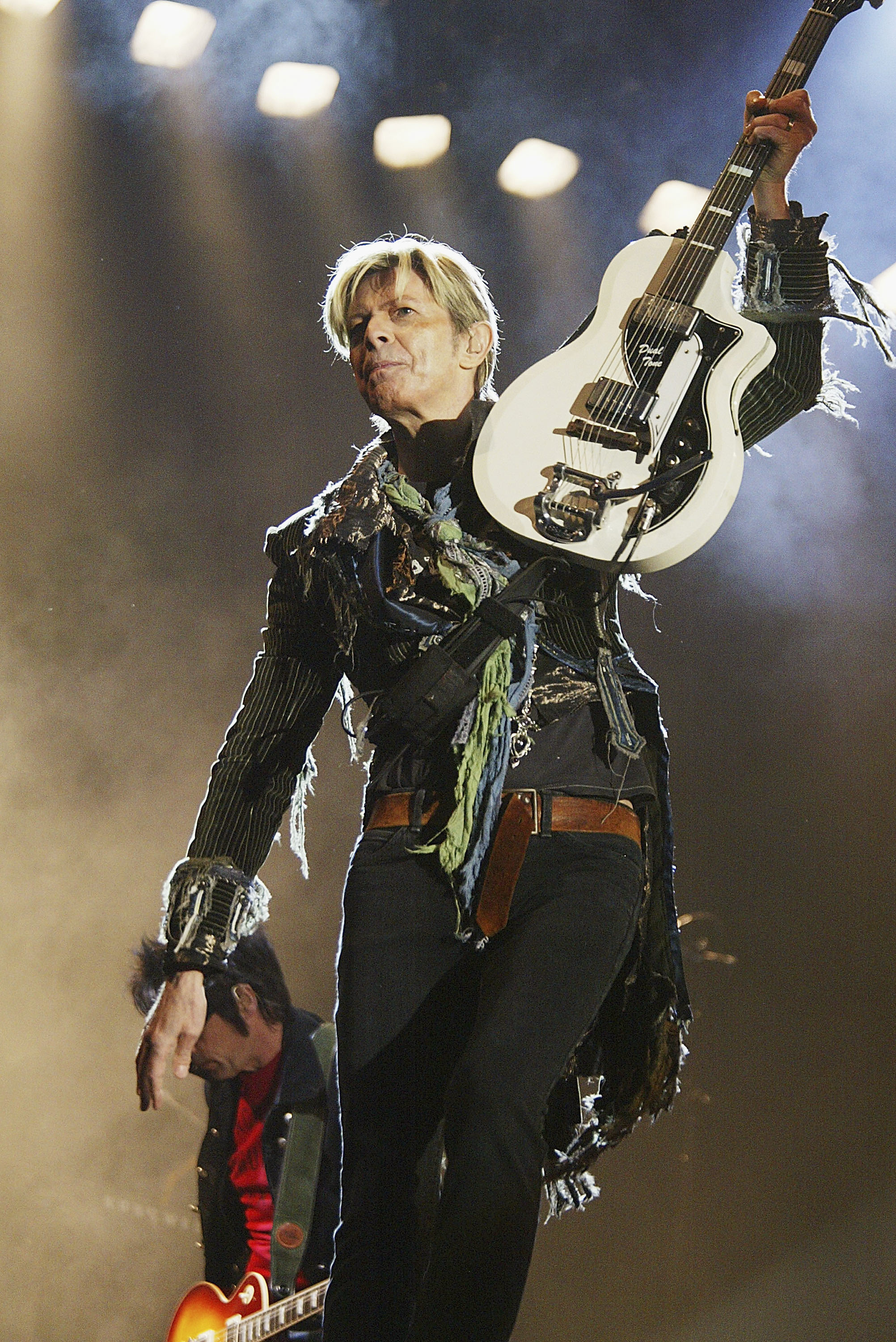 David Bowie, on stage, holds a white guitar above his head. He is wearing a fringed jacket and belted pants. Another musician is partially in the background