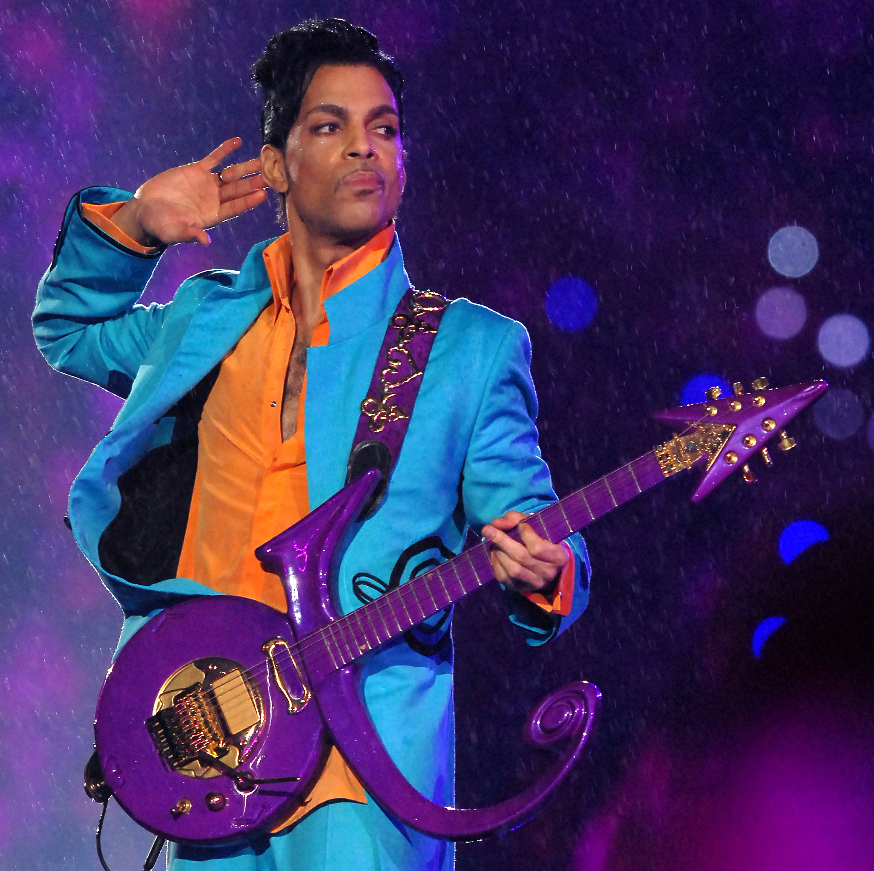 Prince performs on stage, holding a distinctive purple guitar and wearing a vibrant suit with an open collar shirt. He raises one hand to his ear