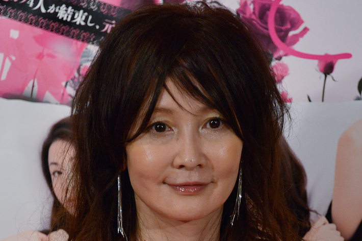 女性がピンクのノースリーブドレスを着て、カメラに向かって微笑んでいる。背景に花や人物のイメージがある。