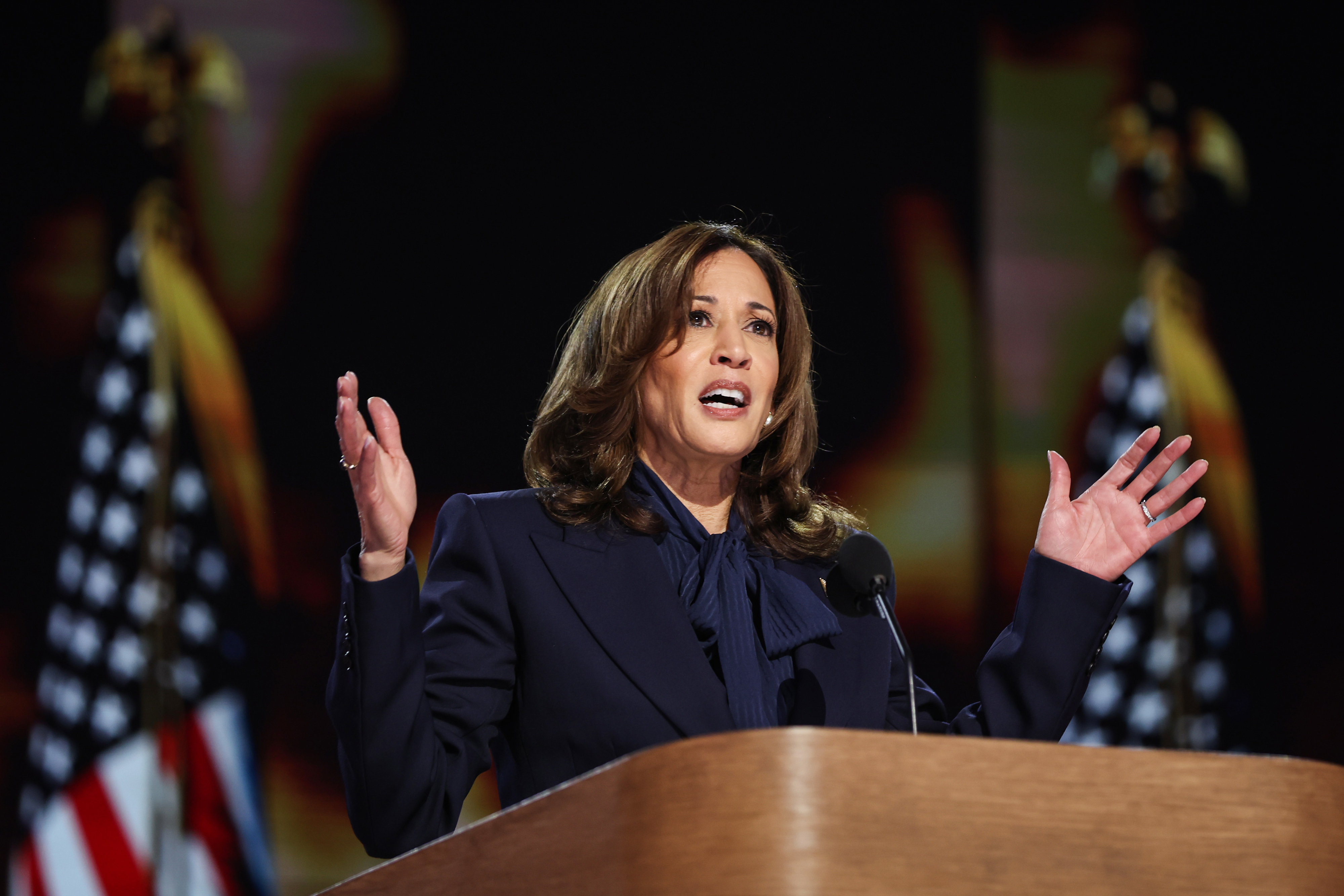 Kamala Harris speaking passionately at a podium, gesturing with both hands, with American flags in the background